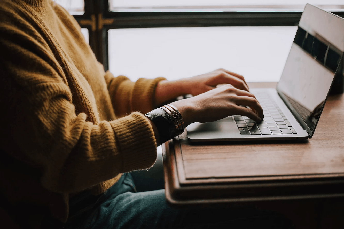 Person in gelbem Pullover tippt auf Laptop an Holzschreibtisch. Sonnenlicht vom Fenster im Hintergrund. Fokussierte, ruhige Stimmung.