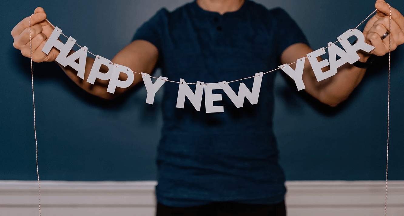 Person hält ein “HAPPY NEW YEAR” Banner vor blauem Hintergrund. Banner in Weiß, Person trägt ein dunkelblaues Shirt. Feierliche Stimmung.