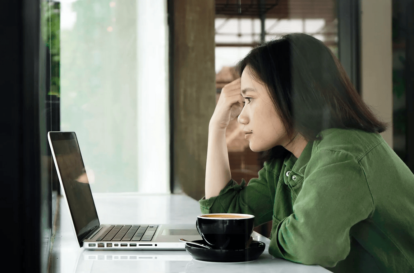 Frau in grünem Hemd blickt konzentriert auf Laptop in Café. Schwarze Tasse im Vordergrund, Fenster im Hintergrund. Ruhige Stimmung.